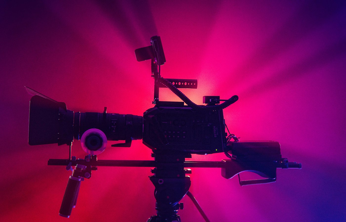 A cinema camera silhouetted against a wall lit in magenta and violet.