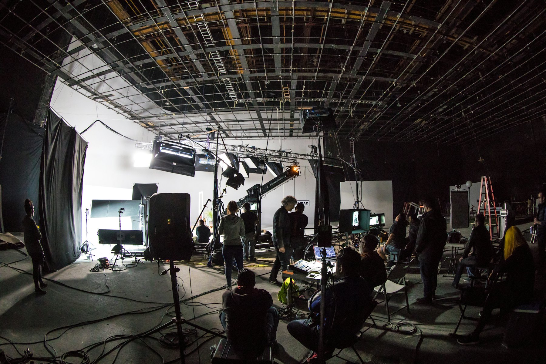 A film crew at work on a large soundstage, with lighting rigs overhead and cameras set up on the floor.