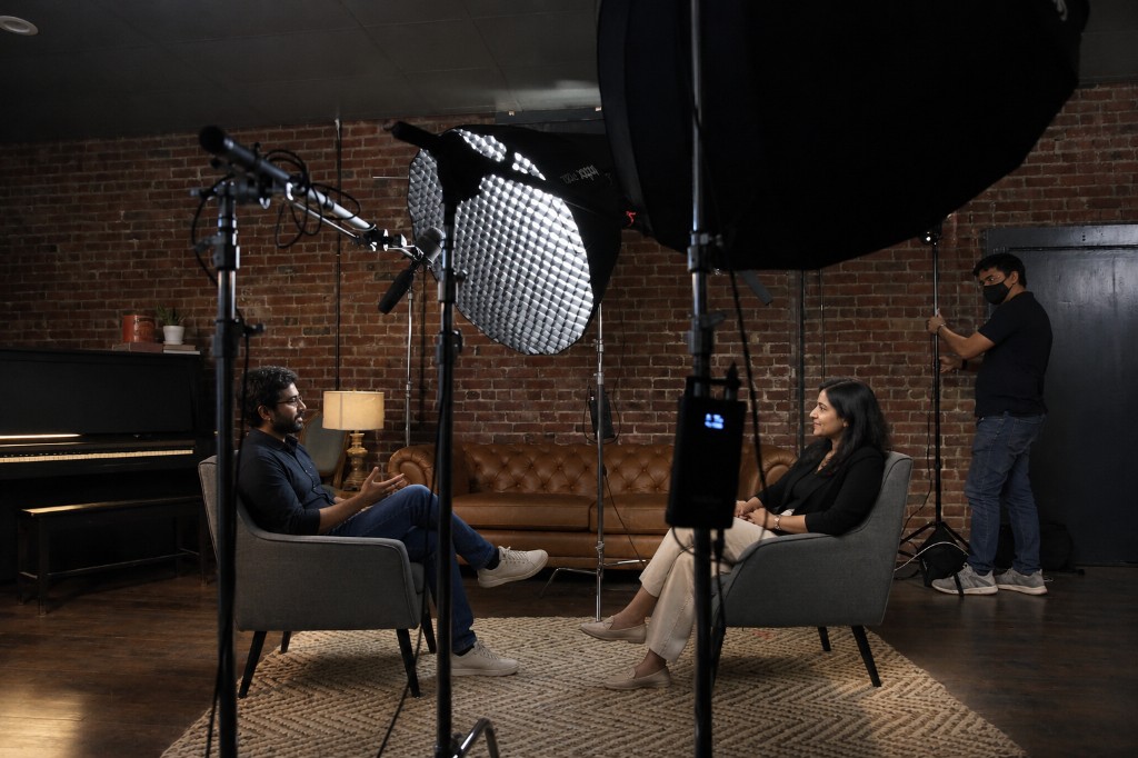 Two people seated for an interview in a small studio, with soft lights and a brick wall behind them.