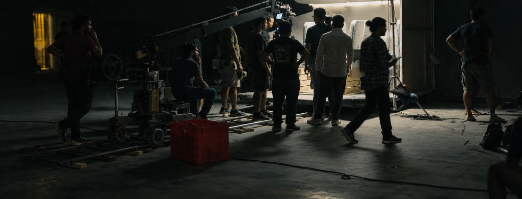 A film crew on a darkened stage floor, gathered around a camera crane and a lit set.